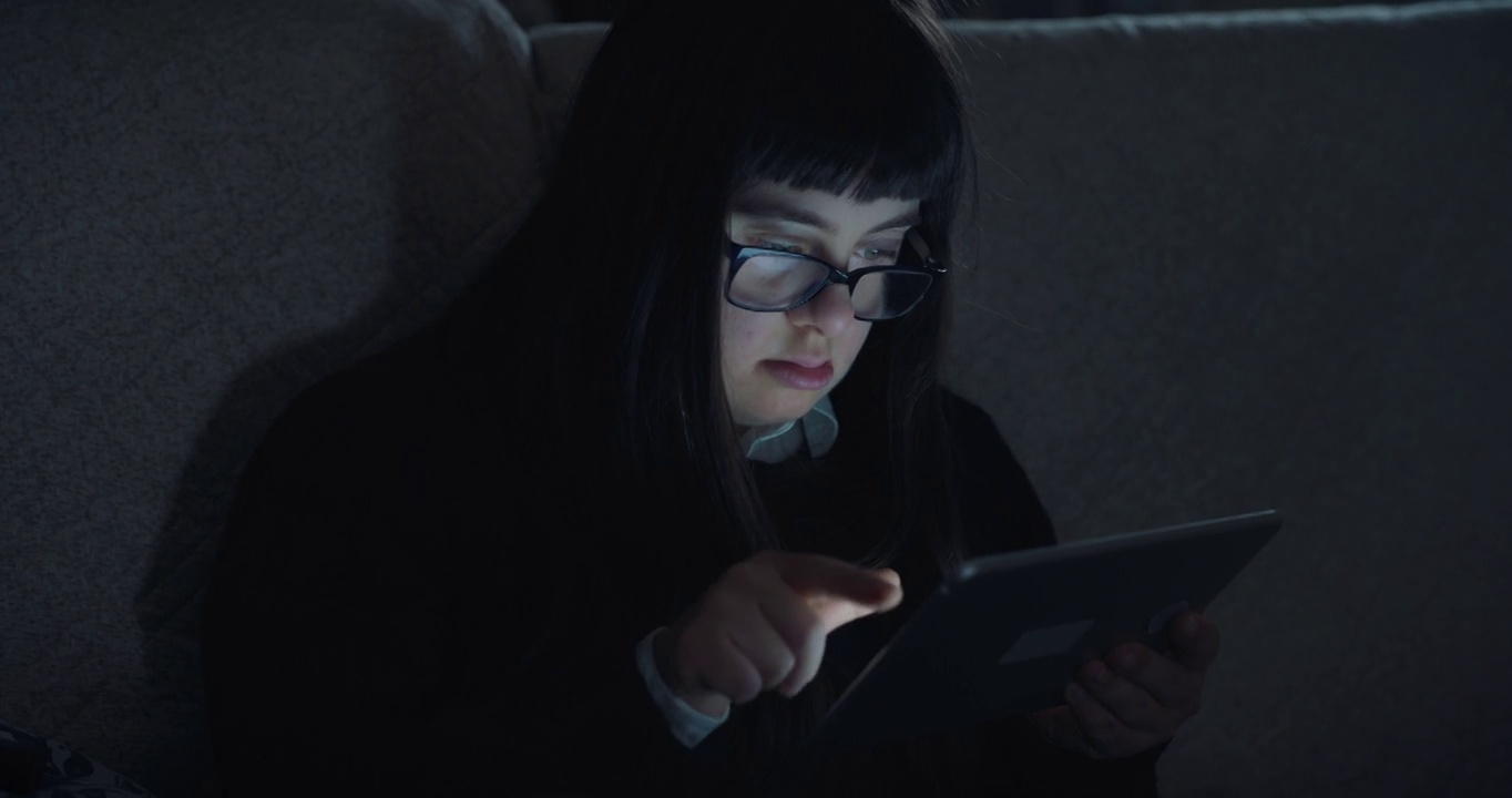 一个患有唐氏综合症的十几岁女孩晚上坐在家里的沙发上用平板电脑娱乐。技术概念，连接，残障，残障，在线，沟通