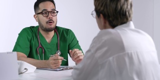 身着外科制服的年轻亚洲男医生微笑着向在检查室使用笔记本电脑的女病人讲解。