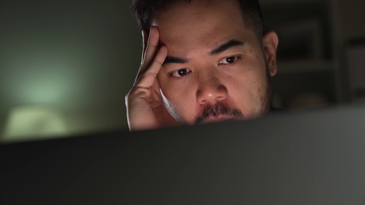 亚洲男人在家里使用电脑工作到很晚时，感觉不开心，有压力和疲倦。