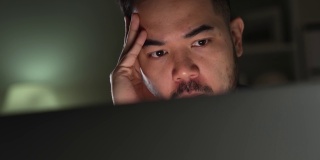 亚洲男人在家里使用电脑工作到很晚时，感觉不开心，有压力和疲倦。
