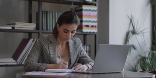 女商人在笔记本上写东西。创业女企业家学生在工作场所靠近电脑的地方学习写笔记。一位女士在桌上的空白笔记本上手写着什么。