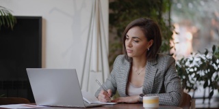女商人在笔记本上写东西。创业女企业家学生在工作场所靠近电脑的地方学习写笔记。一位女士在桌上的空白笔记本上手写着什么。