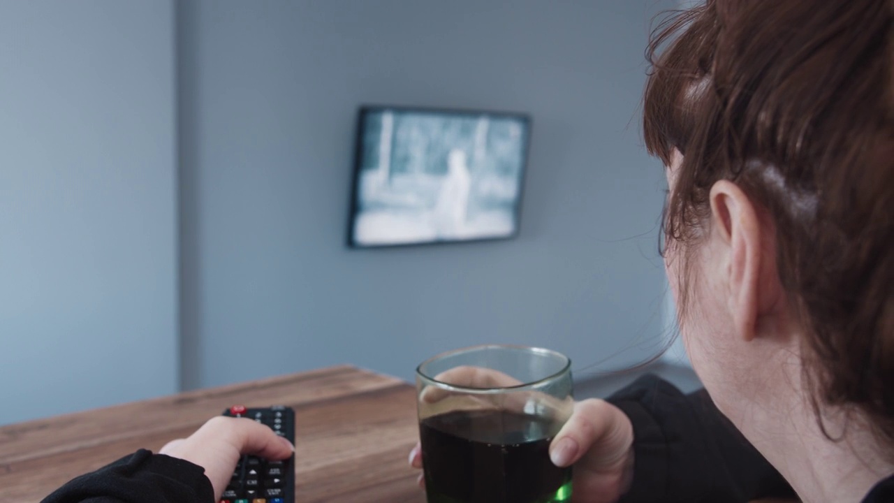 一个女人正在切换电视频道，选择一个频道，喝着苏打水