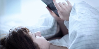 漂亮的黑发女人早上把头放在枕头上，在床上用电话，读一些信息，读新闻