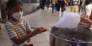 一个亚洲小女孩在街头夜市用特殊的机器制作棉花糖。孩子们拿着一根棉花糖棒，棉花糖棒上缠绕着细长的线。