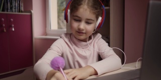 远程学习。用笔记本电脑在线学习的女生在抄写本上做笔记。学生戴耳机观看网络视频课程。白种人女孩戴耳机学习在线课程