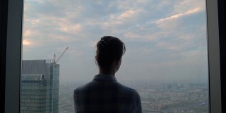 透过摩天大楼的窗户看城市风景的沉思的女人的后视图