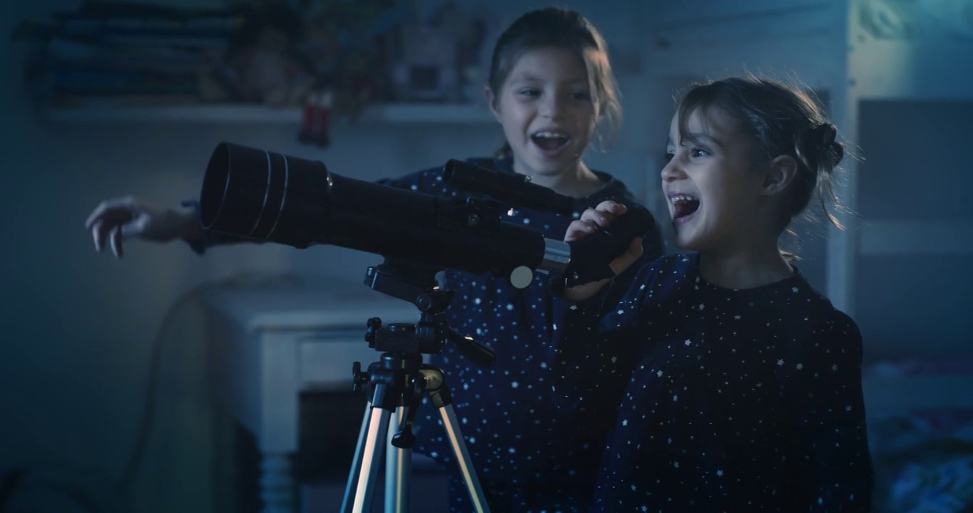 在夜晚拍摄的电影中，穿着缀满星星的睡衣的两个快乐的小女孩正在通过望远镜观看黑暗的天空中坠落的星星。概念:探索、教育、童年