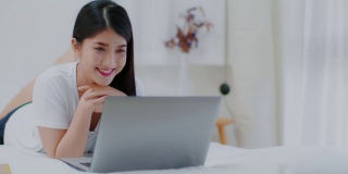 亚洲年轻女性在电脑上打字放松，看社交媒体上的热门视频，和朋友在卧室聊天。有吸引力的女性躺在床上玩和使用笔记本电脑应用程序。