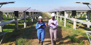 非洲投资者和亚洲中年女工程师身穿蓝色防护服，戴着安全眼镜、头盔在太阳能农场边走边谈，正面高角度观看