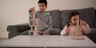 可爱的欧洲孩子一起玩棋盘游戏。一个英俊的学龄男孩，用木砖砌成房子，坐在沙发上，旁边是他的小妹妹，她的塔倒塌了，她很难过