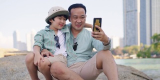 一位亚洲父亲和他快乐的儿子用智能手机自拍，一起在城市附近的岩石岸边度过美好时光