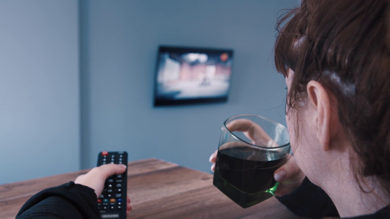 一个女人正在切换电视频道，选择一个频道，喝着苏打水