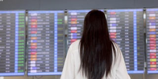 在国际机场的候机楼里，一个留着黑色长发的女商人正拿着泰国护照，看着信息板，查看她的航班和背景模糊的航班时间表。