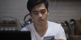 一名谨慎的亚洲男子在夜间在家的餐厅里用信用卡用笔记本电脑在线支付税款的特写镜头