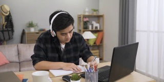 勤奋的亚洲男学生戴着耳机，在家里明亮的客厅里用笔记本电脑上在线课程，记笔记。