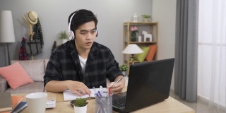 一名勤劳的中国年轻人戴着耳机，一边在家里用笔记本电脑在线学习语言课程，一边在笔记本上写作和练习口语。