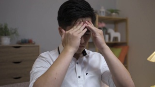 头部照片，一个疲惫的亚洲商人按摩他的头部与痛苦的脸，而遭受严重的偏头痛在家里的客厅。视频素材模板下载