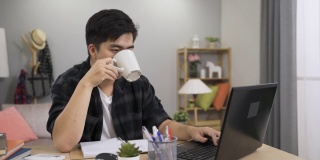 一名亚洲男性自由职业工人一边喝茶，一边在家里的电脑上继续工作，一边点头。