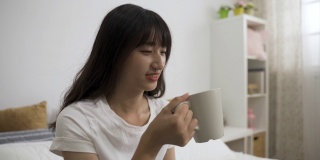 阳光明媚的早晨，一位无忧无虑的韩国女性在家里的卧室里，面带满意地喝着美味的咖啡。