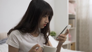 一名亚裔女孩在家里的卧室里端着茶杯，在智能手机屏幕上滚动着查看令人惊讶的网络新闻。视频素材模板下载