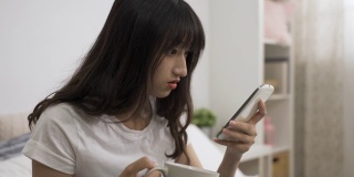 一名亚裔女孩在家里的卧室里端着茶杯，在智能手机屏幕上滚动着查看令人惊讶的网络新闻。