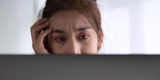 年轻的亚洲妇女坐在看起来筋疲力尽，在使用笔记本电脑在家庭办公室工作。忧心忡忡的亚洲妇女看着笔记本电脑，对电脑问题感到沮丧