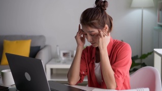 年轻的亚洲妇女坐在眼睛闭从疲惫和疲倦在工作在家庭办公室使用笔记本电脑。劳累过度的亚洲女学生工作人员，在电脑工作后因疲劳而按摩眼睛和太阳穴视频素材模板下载