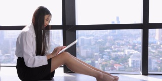 中国一名留着长发的年轻女子，在早上上班前独自坐在窗前，用平板电脑发邮件。