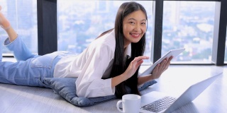 一名年轻的亚洲女性，双腿翘起，躺在地板上，在家用笔记本电脑或平板电脑观看在线数字课程，帮助员工提高技能。商学院为成人提供课程教育。