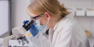 穿着个人防护装备的实验室女研究人员通过显微镜观察并在笔记本电脑上记录结果