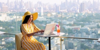 优雅的亚洲女人穿着黄色的裙子，戴着草黄色的帽子，看着城市风景和网上购物的笔记本电脑与鸡尾酒，平板电脑，智能手机在桌子上。
