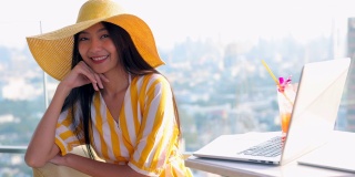 漂亮的亚洲女人多种族美丽优雅的年轻女人在黄色的裙子戴着稻草黄色的帽子看着相机与笔记本电脑和鸡尾酒在桌子上。