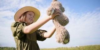 小女孩抱着一只泰迪熊。绿色连衣裙，草帽。蓝天碧草。