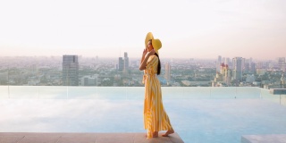 美丽的女人走在池边，穿着黄色的长裙和黄色的帽子。女人们漫步在无边的游泳池旁，伴着夕阳的城市风光。