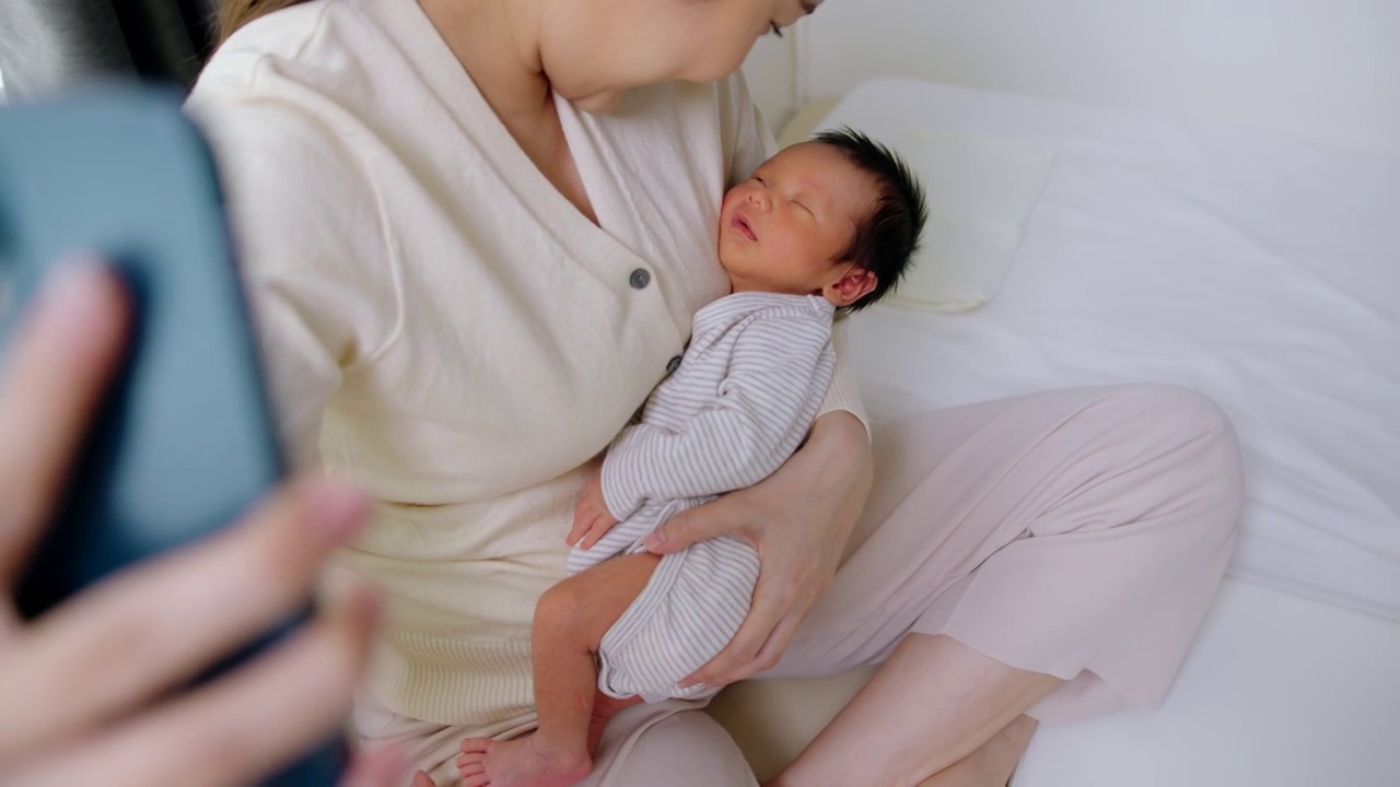 美丽的母亲抱着可爱的新生儿用手机视频通话。技术