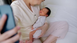 美丽的母亲抱着可爱的新生儿用手机视频通话。技术视频素材模板下载