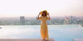 美丽的照片，亚洲妇女在一个黄色的长裙和黄色的大帽子走在池边在多彩的日落。女孩享受暑假与美丽的日落和城市的光线。