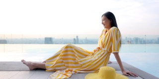 一名年轻的中国女子穿着黄色连衣裙，戴着帽子，坐在摩天大楼的无边泳池边上。女人享受城市的风景，日落时的自然，酒店的屋顶