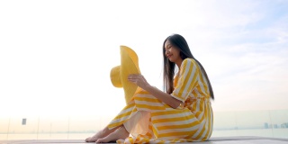 美丽的亚洲妇女在黄色的衣服和黄色的帽子坐在池边，看着相机和放松日落在工作日休息或完成工作后。
