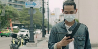 2019冠状病毒病(covid-19)大流行期间，一名亚洲男子戴着医用口罩，在城市街道上行走时使用智能手机。