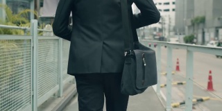 跟踪镜头:一名亚洲商人穿着休闲装，提着肩包走在大街上上班。