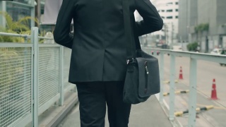 跟踪镜头:一名亚洲商人穿着休闲装，提着肩包走在大街上上班。视频素材模板下载