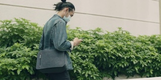 2019冠状病毒病(covid-19)大流行期间，一名亚洲男子戴着医用口罩，在城市街道上行走时使用智能手机。