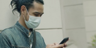 在covid-19大流行期间，一名年轻的亚洲男子戴着医用口罩，站在城市的街道上使用智能手机。