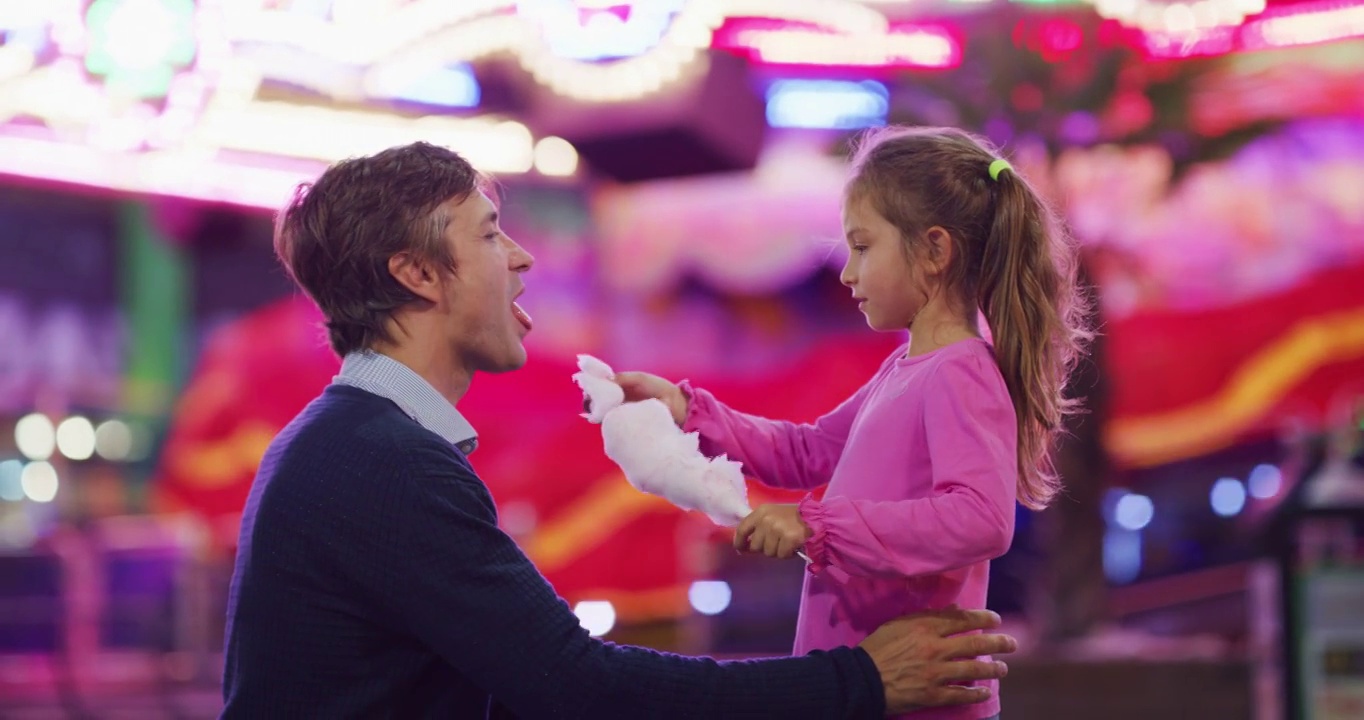 真实的照片，一个快乐的父亲和他的小女儿正在开心地吃棉花糖在游乐园与月亮公园的灯光在晚上。