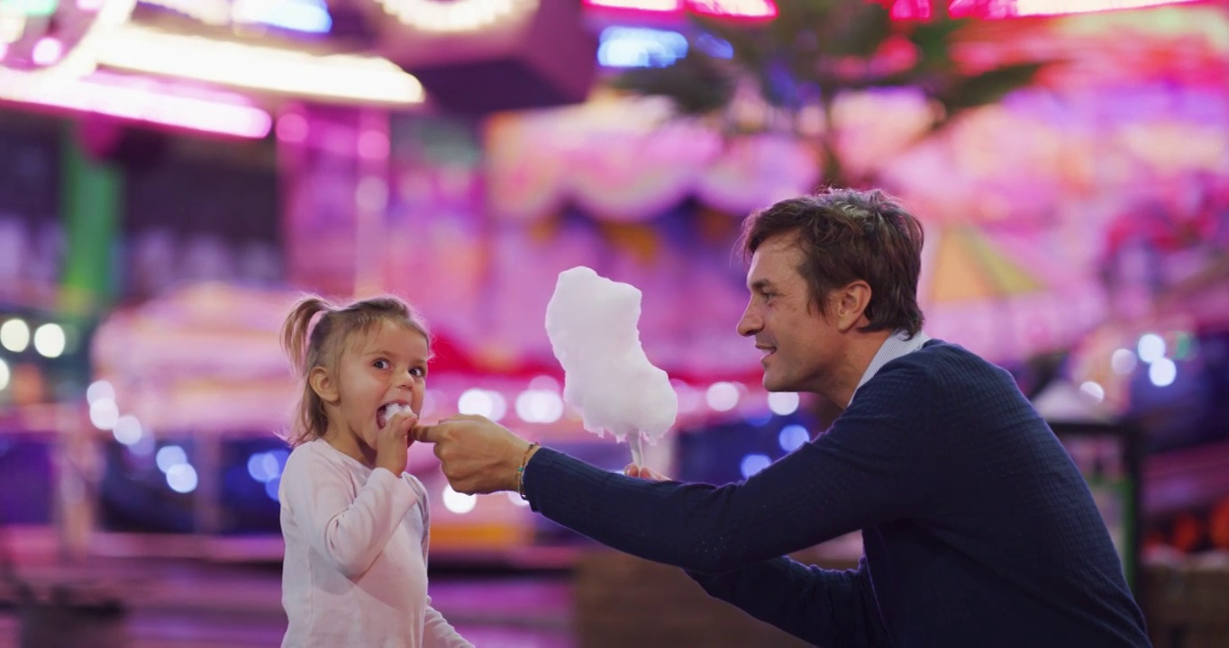 真实的照片，一个快乐的父亲和他的小女儿正在开心地吃棉花糖在游乐园与月亮公园的灯光在晚上。