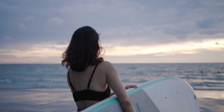 亚洲妇女持有一个sup板和划桨和行走在海滩上。背景是大海和日落。后视图。夏天冲浪。