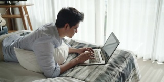 年轻的亚洲商人躺在床上，用笔记本电脑在度假胜地附近的房间在暑假假期旅行。