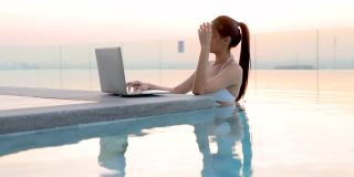 年轻的中国女子穿着白色比基尼在游泳池放松。女人享受工作，网上购物，检查社交网络，阅读新闻在舒适的笔记本电脑在早晨。